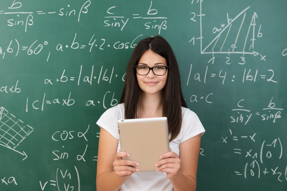 Clever attractive young female college student wearing glasses and holding a tablet computer in her hands standing in front of a chalkboard covered in equations in maths class smiling at the camera Clever attractive young female college student wearing glasses and holding a tablet computer in her hands standing in front of a chalkboard covered in equations in maths class smiling at the camera